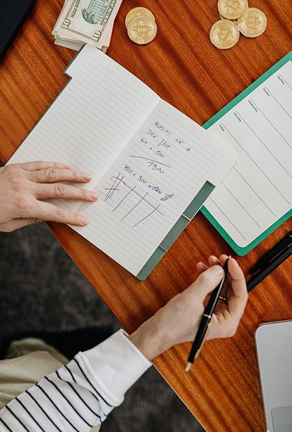 Overhead view of hands writing calculations in a notebook with cash and coins on a desk