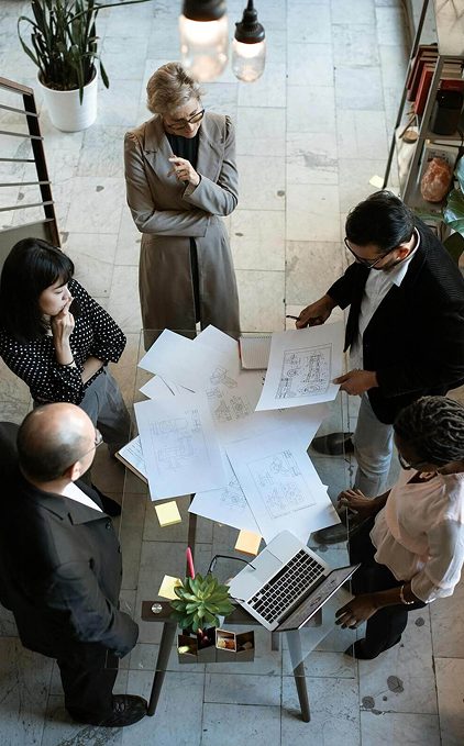 Overhead view of a team standing around a table reviewing printed plans and documents together during a planning meeting.