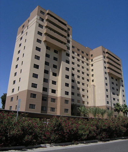 Tall multi-story residential building with tan and brown stucco exterior, flowering shrubs along the sidewalk in front.