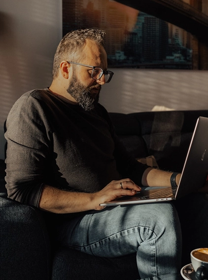 Bearded man with glasses sitting on a couch working on a laptop with a coffee cup nearby.
