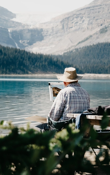 Man in a wide-brimmed hat sitting at a picnic table looking out over a mountain lake with glaciers in the distance.