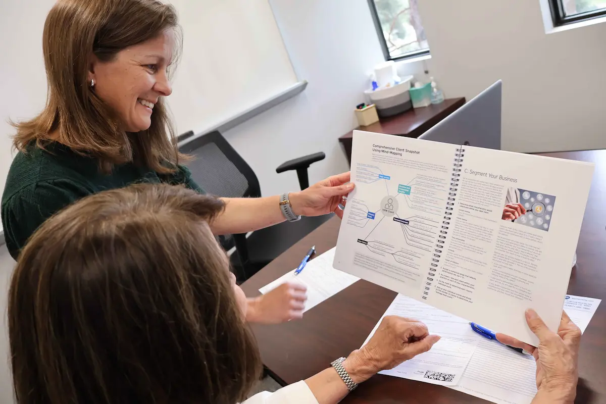 Smiling financial advisor and a client holding open a spiral-bound planning guide showing a client snapshot mind map, with forms and pens on the desk in front of them.