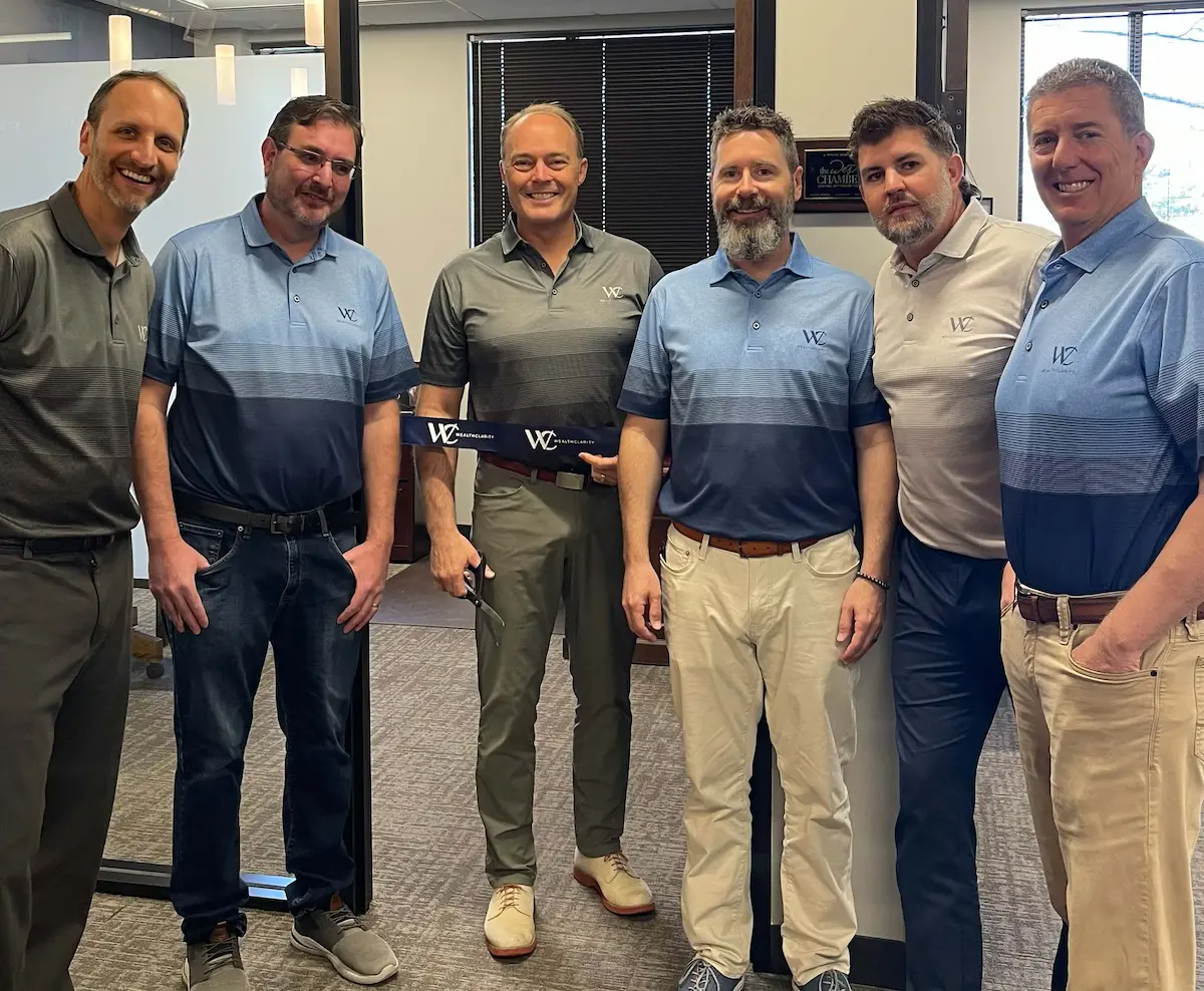 Six members of the WealthClarity team in matching logo polo shirts smiling at a ribbon-cutting for the firm's office, with one holding scissors and a WealthClarity-branded ribbon.