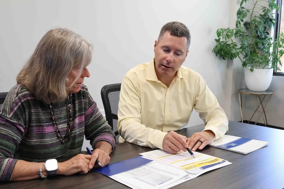 Financial advisor pointing with a pen to highlighted figures on a planning document as a client reviews the materials with him at a conference table.