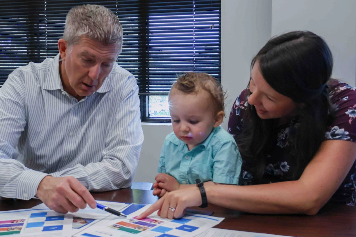 Financial advisor pointing to a planning document with a pen while a mother, holding her toddler on her lap, reviews the materials with him at a conference table.